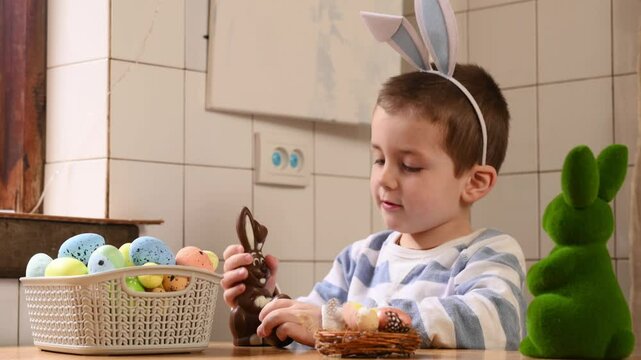 Cute little Boy with bunny ears plays with colored painted eggs, closes eyes with toy, plays hide and seek. Egg hunt, traditional catholic orthodox easter.  Little boy celebrate Easter at home