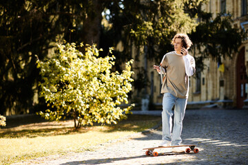 Fototapeta premium Stylish young man skateboarding through a sunlit path in the park while on the phone