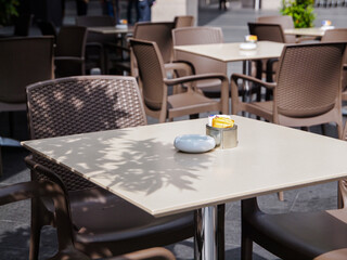 A table seating layout of a cafe outdoor with plastic brown chairs and off white beach table top, an ashtray and a metal can with sugar and cream packets.