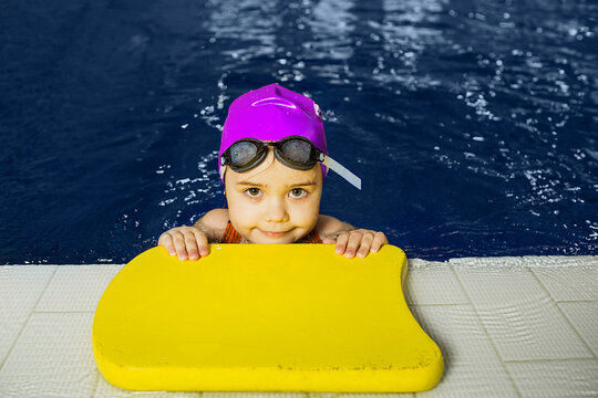 A child is practicing swimming techniques at an indoor pool, using a flotation device while wearing a cap and goggles