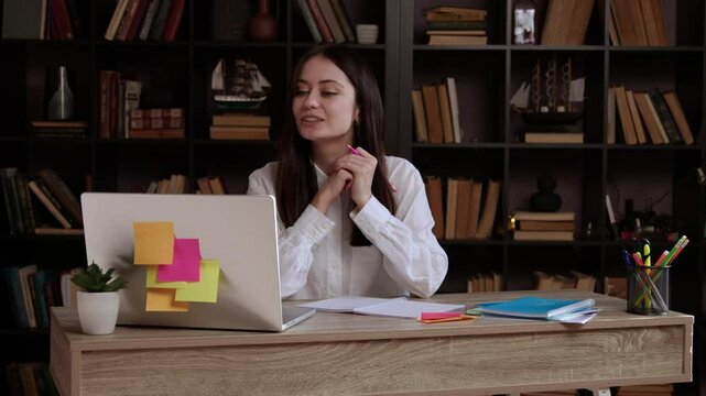 Smiling female student at an office table and in a modern library presenting her online bachelor's thesis in front of the computer and the licensing committee. Student explains the completed project