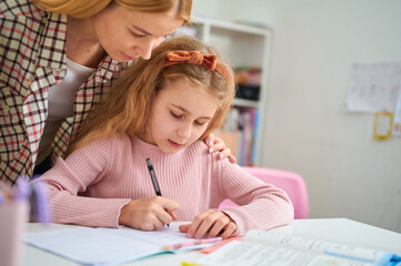 Mother helping daughter with homework at desk,  looking to workbook and explaining tasks in a...