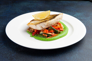 Cod steak on a bed of vegetables in a plate on a dark background.