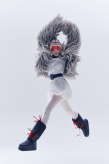 Young woman mid-air in oversized shaggy fur coat, ski goggles, and moon boots against white studio...