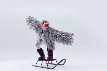 Young woman balancing on sled in oversized shaggy fur coat, ski goggles, and moon boots against...