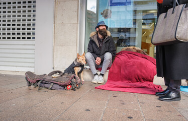 Homeless man with dog sitting on sidewalk and looking up while elegant woman is giving him alms