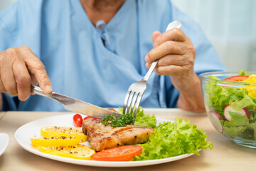 Asian senior woman patient eating pork chop stake and vegetable salad for healthy food in hospital.