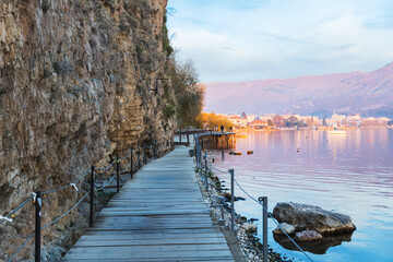 Wooden walkway by a rocky cliff along the serene Lake Ohrid. Calm waters reflect the distant town...