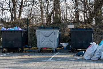 Multiple trash bins are filled with garbage in a parking area, surrounded by scattered litter and...