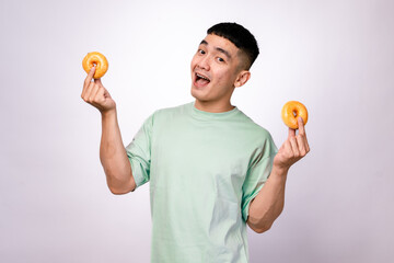 An Asian man in a mint green shirt is holding two glazed donuts on his fingertips while smiling widely. He is looking happy and playful against a plain white background.