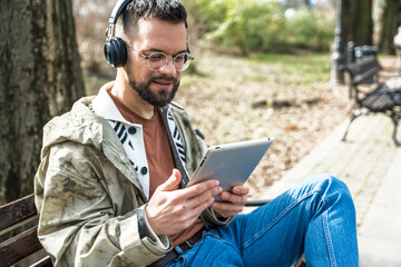 Obraz premium Young hipster businessman with cool attitude sitting in the park having video call on digital tablet. Expatriate business person standing out of the crowd, intelligent design professional occupation.