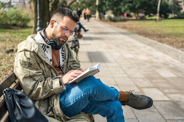 Young curious religious man, reading a book about religion, while sitting in the park on bench. Finding God male, happy and hungry for knowledge about faith. Christian or muslim read Bible or Quran.