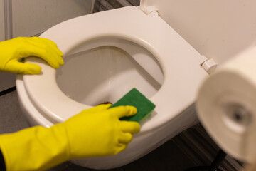 Hands in yellow rubber glove with sponge cleaning dirty toilet bowl.