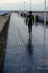 A barefoot surfer in a wetsuit walks along a wet promenade near the ocean, carrying a surfboard. The reflections on the wet pavement create a striking visual effect