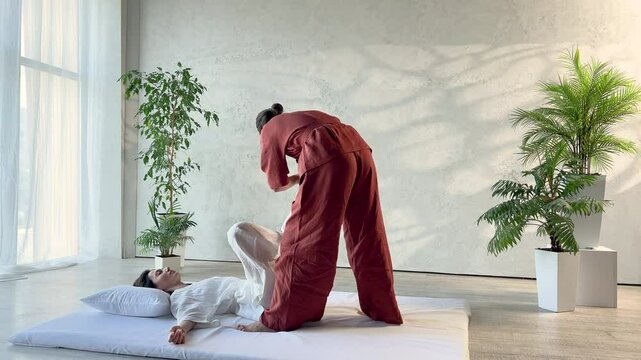 Masseuse making thai yoga massage. Woman therapist doing traditional massage treatment, stretching female patient's body. Client lying on mat, enjoying hand massage in calm atmosphere.