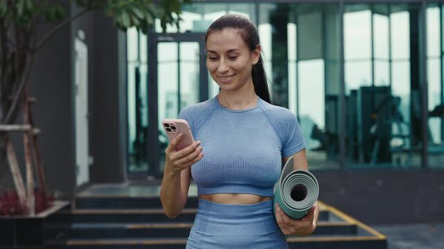 Fit woman in athletic attire holds yoga mat using at phone in gym. Happy female communicates on social media walking to training. Fitness blogger makes content for personal page