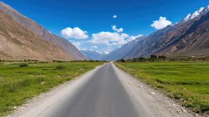 Fototapeta premium Scenic mountain road through a lush green valley under a vibrant blue sky with snow-capped peaks in the distance.