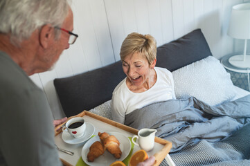 Elderly man serving breakfast in bed to his smiling wife. A cozy morning moment with coffee, croissants, and fresh orange juice, symbolizing love, care, and a happy senior lifestyle.