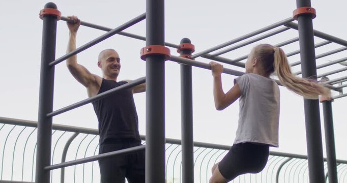 Father and blonde daughter do pull-ups on horizontal bars. Junior schoolgirl looks at dad as role model dropping down after attempting exercise
