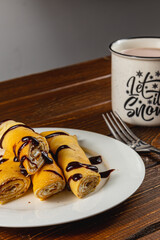 Rolled pancakes with chocolate drizzle and a cozy drink on wood