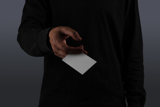 a person dressed in a black long-sleeve shirt, extending their hand forward while holding a dark, sleek business card. Isolated blank mockup surface to place your design. 