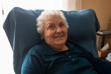 portrait of a woman in her eighties smiling and looking at the camera in an armchair in her house,...
