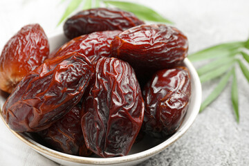 Many tasty dried dates in bowl and leaves on gray textured table, closeup