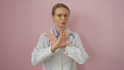 Doctor woman with blonde hair making a stop gesture over a pink background looking serious wearing a stethoscope and glasses indicating caution or prevention