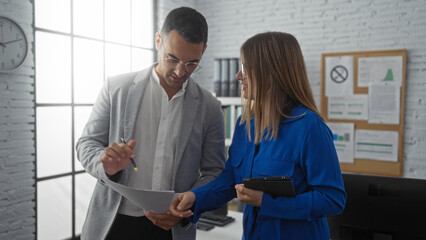 Obraz premium Man and woman discussing documents in modern office environment, with large windows and white brick walls, conveying a collaborative professional atmosphere.