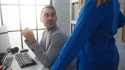 Man sitting at desk looking at woman in office setting with computer and bookshelves, indicating a business interaction in a professional workplace environment.