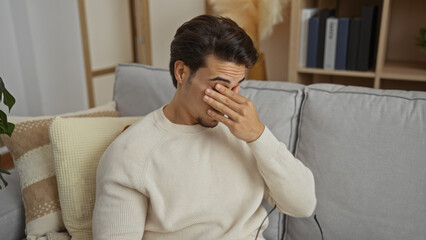 Young man sitting on a sofa in a living room covering his eyes with his hand, suggesting a moment...