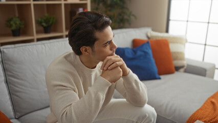 Young hispanic man in cozy living room sitting on sofa with thoughtful expression amid modern decor...