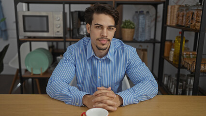Handsome hispanic man in blue shirt sitting at office table with modern decor in workplace interior surrounded by shelves and kitchenware, looking confidently at camera.