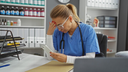 Nurse wearing blue scrubs and stethoscope checks phone looking stressed in a clinic's organized...