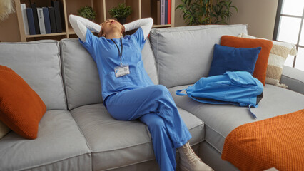 Woman relaxing on a sofa at home wearing blue scrubs with a backpack beside her in a cozy living room setting with cushions and bookshelves showing calm and relaxation.