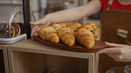 Woman arranging croissants in a cozy bakery shop with wooden shelves surrounded by fresh pastries under warm indoor lighting