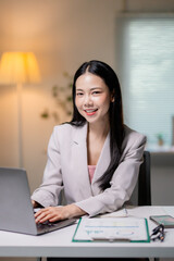 Professional Asian woman working on laptop, smiling at desk in contemporary workspace