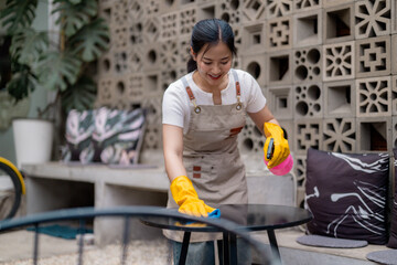 Smiling waitress wearing apron and gloves cleaning table with disinfectant spray and cloth in outdoor cafe