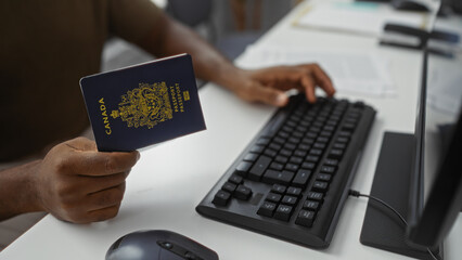 Man holding canadian passport at office desk using computer keyboard and mouse, representing professional work environment with focus on international travel.