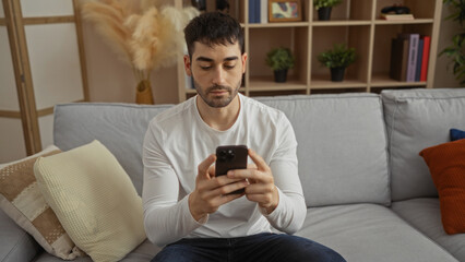 Fototapeta premium Young man using smartphone indoors on a comfortable sofa in modern living room suggests relaxation and technology at home.