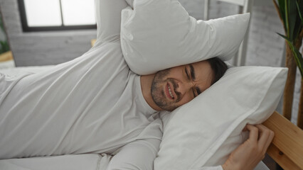 Young man in bedroom lying on bed wearing white shirt with pillow over head appearing to be woken up by noise or discomfort in an indoor setting with modern decor