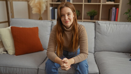 Obraz premium Woman sitting on couch in cozy living room with plants, wearing sweater and jeans, showcasing an inviting and relaxed indoor atmosphere with a contemporary style.