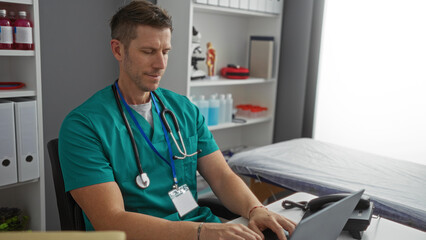 Young man in hospital room wearing scrubs and stethoscope focused on computer work with shelves and medical equipment in the background