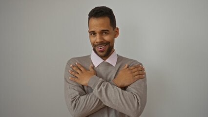 Young hispanic man embraces himself warmly, exuding confidence and charm, set against a plain white background, highlighting his attractive and elegant appearance in a simple yet striking pose.