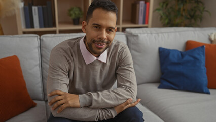 Fototapeta premium Young hispanic man sitting in a cozy living room with stylish decor, exuding confidence and friendliness, surrounded by colorful pillows and bookshelves indoors.
