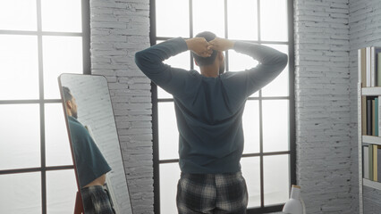 Man standing in bedroom facing mirror with hands on head reflecting on window lit morning indoors