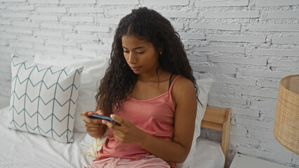 Young woman sitting on a bed in a modern bedroom, using her smartphone with a focused expression,...