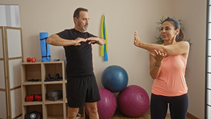 Fototapeta premium Middle-aged hispanic couple exercising in a gym with equipment, showing love and engagement, man and woman stretching together indoors, promoting healthy lifestyle.