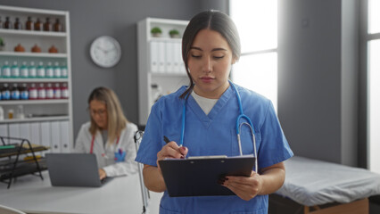 Hispanic female doctors in a clinic room, one writing on a clipboard, the other working on a...