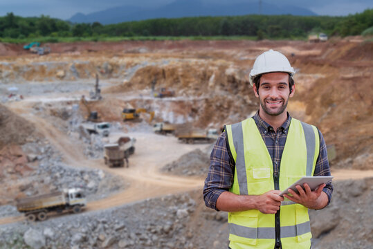 A smiling engineer wearing a hard hat and safety vest holds a tablet at an active mining site with trucks and excavators.